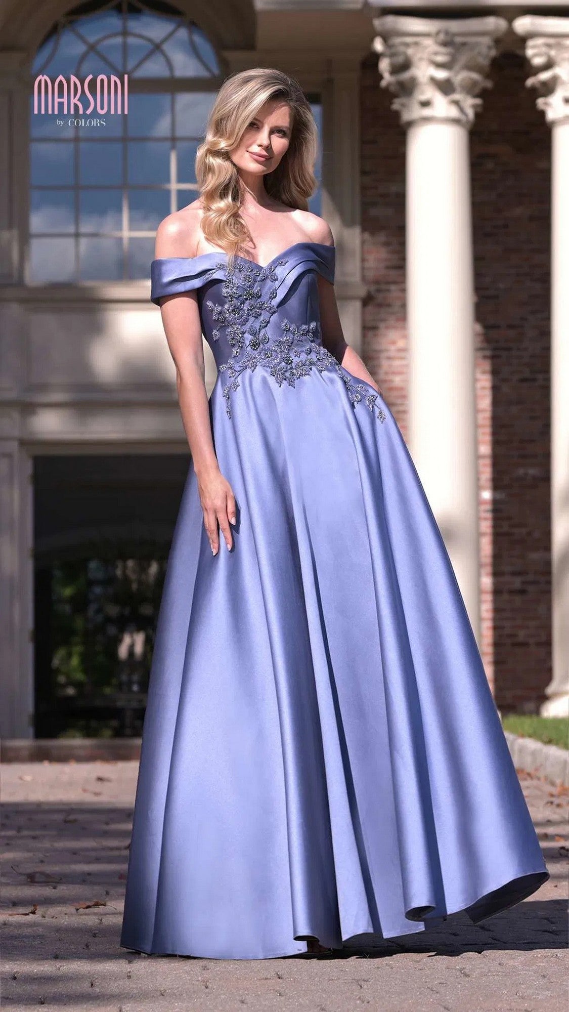 A woman stands outdoors in front of columns, wearing the Marsoni MV1008 long formal dress in floor-length slate blue with an off-the-shoulder design and floral embroidery on the bodice.